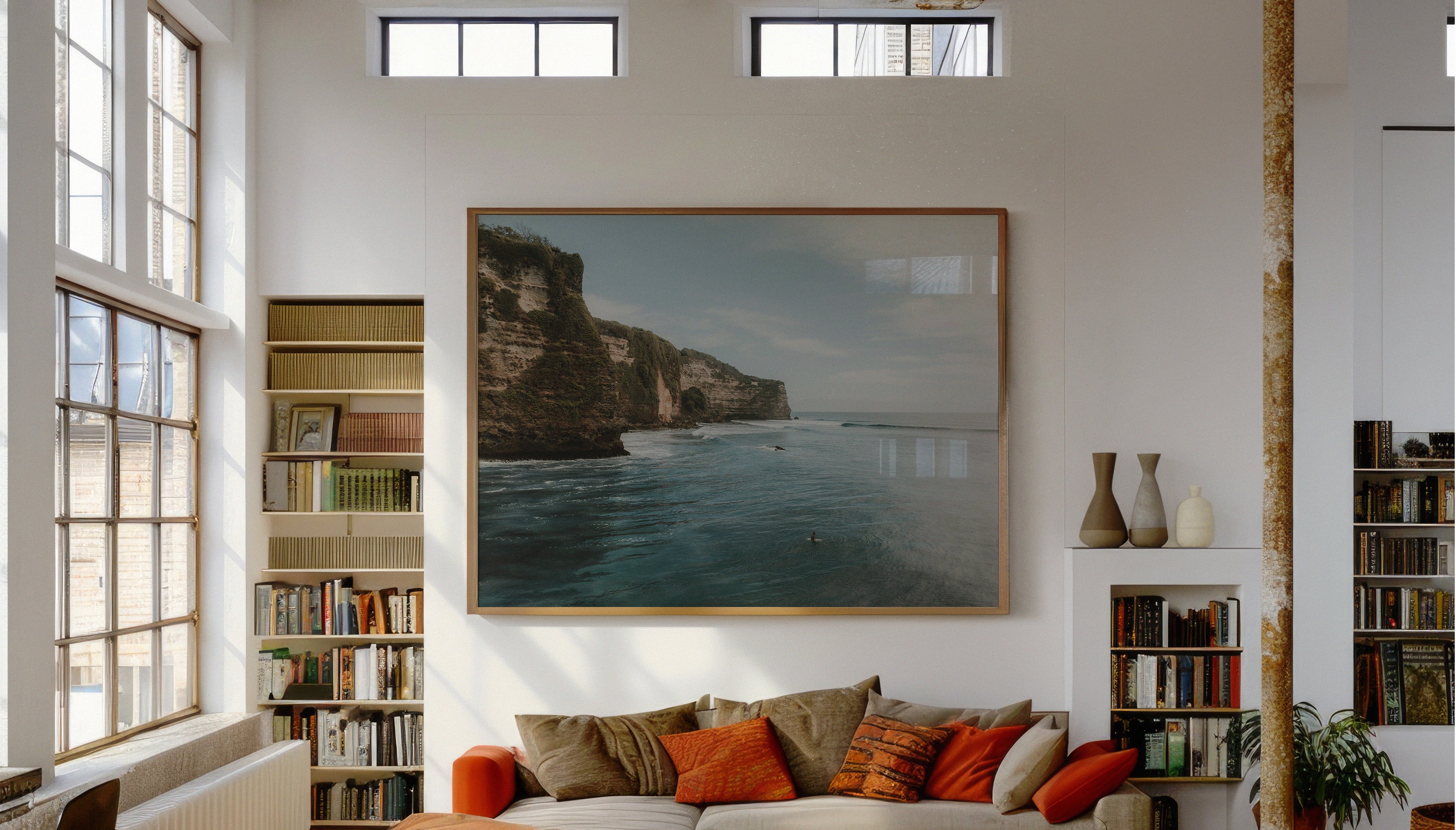 A fine art travel photograph by Sam Morris in a large wooden frame, capturing a surfer sitting in dark turquoise sea below dramatic cliffs covered in wild greenery.