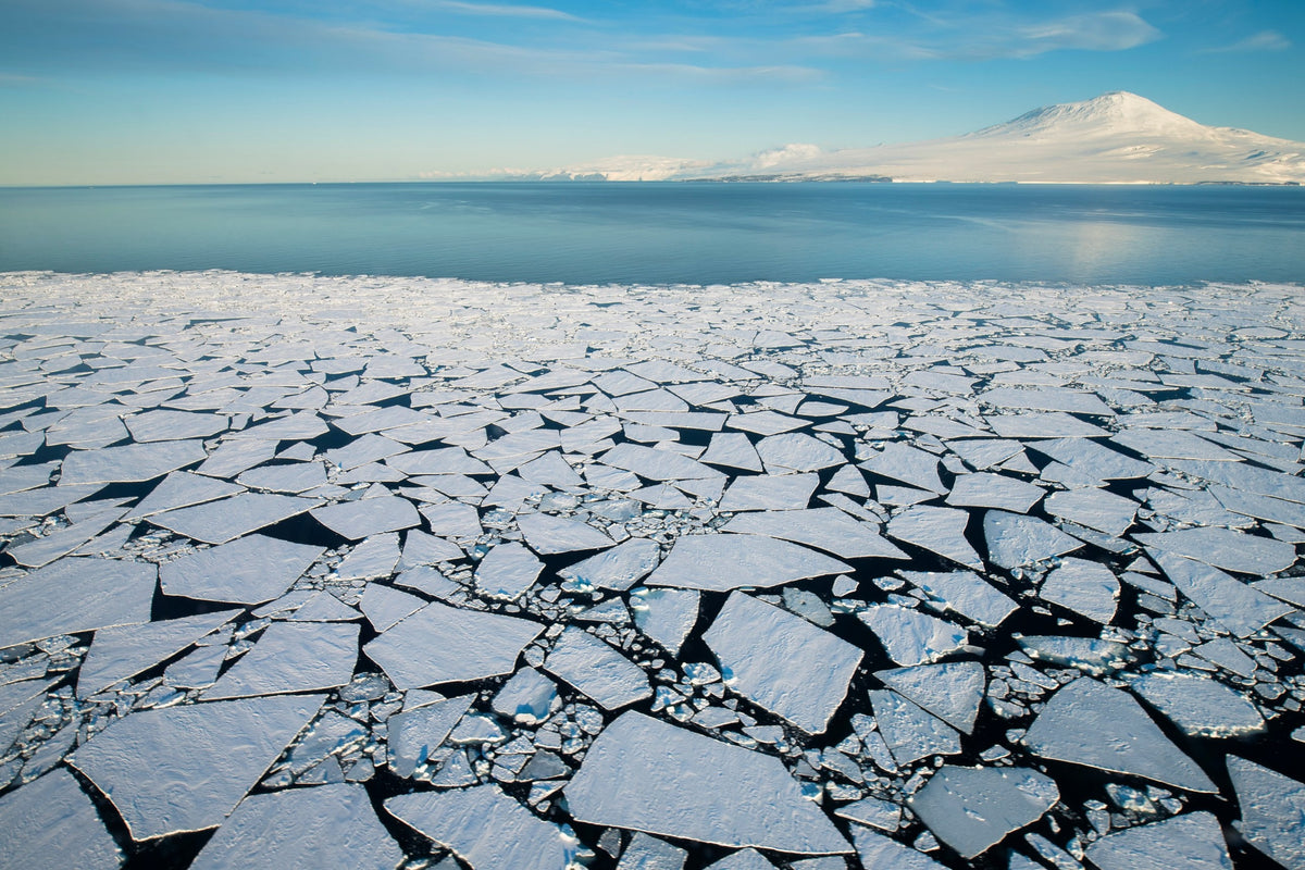 Aerial view fine art print of sea ice broken into white floes across dark water in the Ross Sea, Antarctica, with snow-covered mountains under a blue sky.
